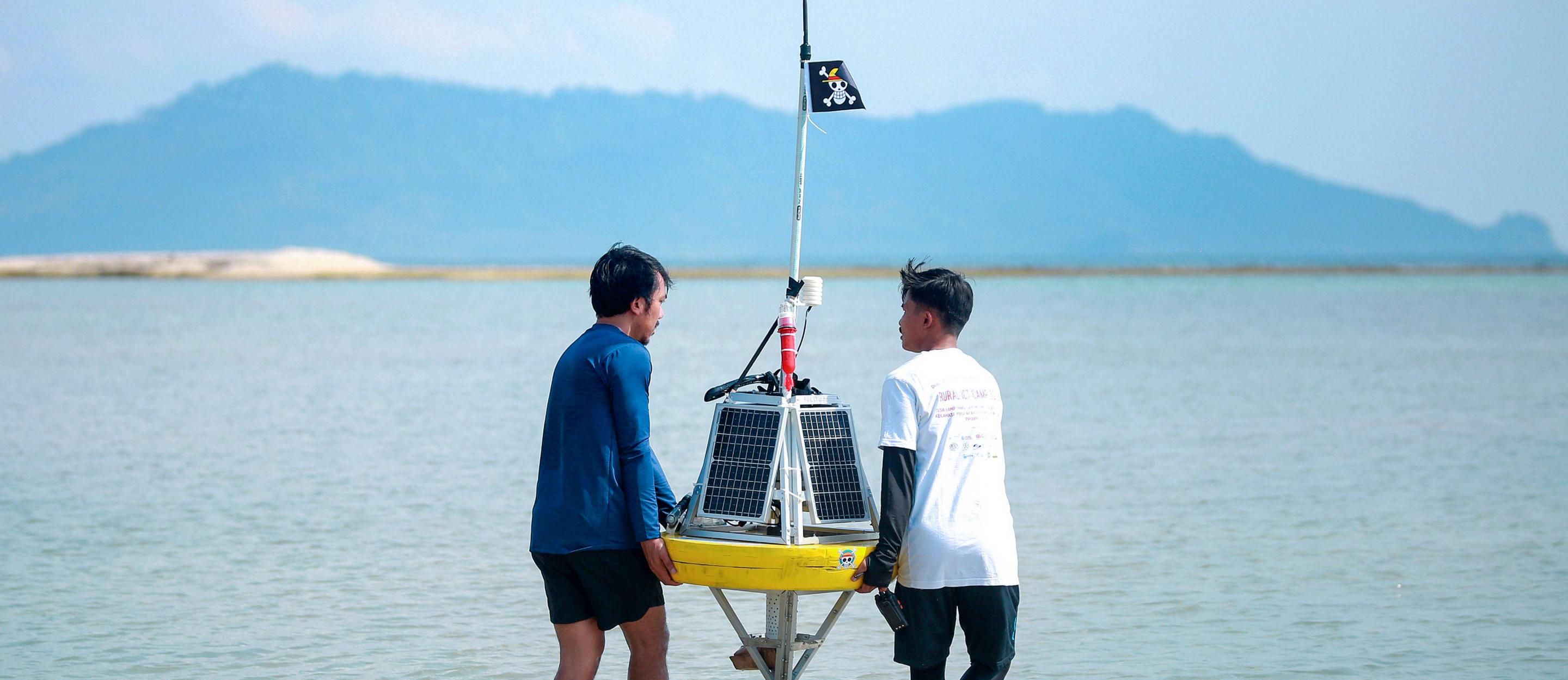  Zwei Männer tragen eine solarbetriebene Messboje durch flaches Wasser, im Hintergrund sind Berge und eine Sandbank zu sehen.