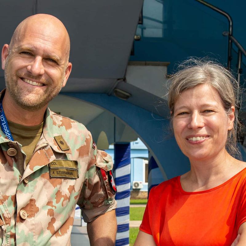 Ein lachender Mann in einer militärischen Tarnuniform mit Namensschild „A. Stirnimann“ steht neben einer Frau in einem roten Shirt vor einer modernen blauen Gebäudestruktur.
